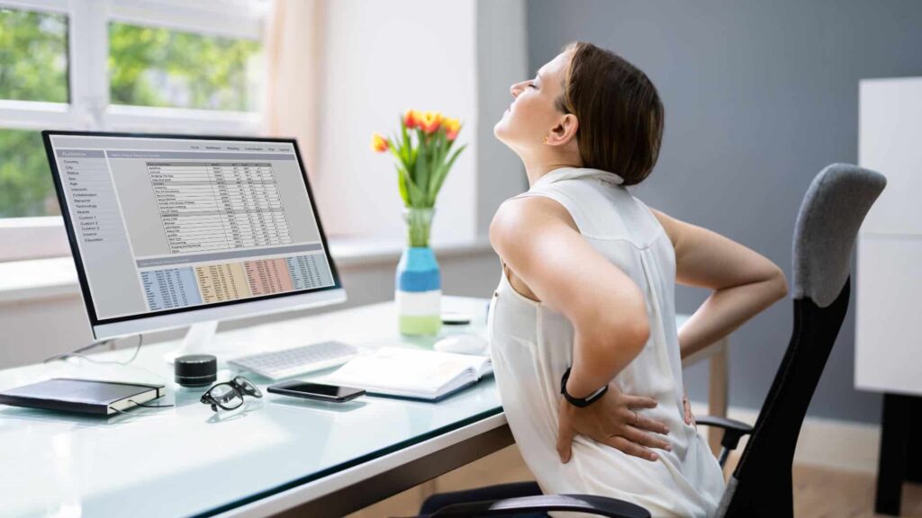woman sitting at a desk with a computer holding her back that is in pain